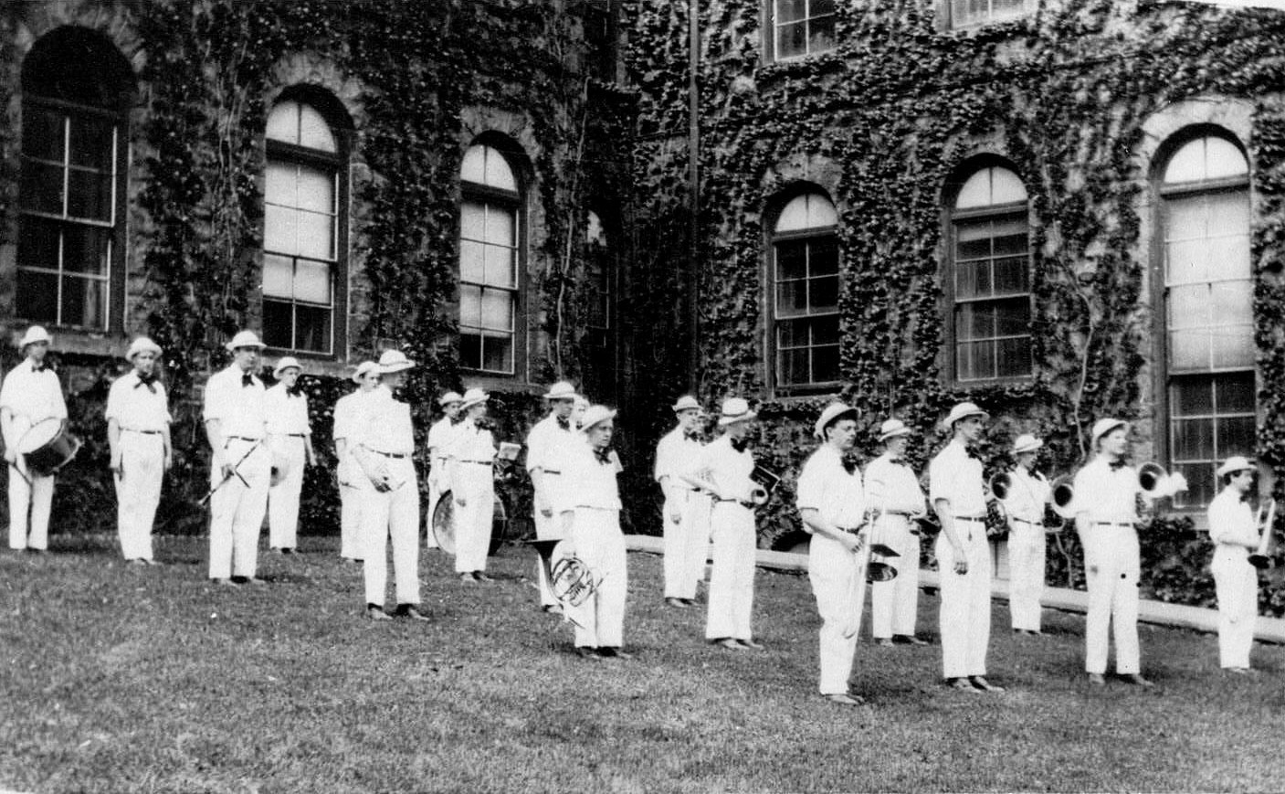 The first Lehigh Band standing in the grass outside a campus building. They are wearing white uniforms and have instruments in hand